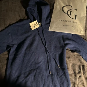 Mörkblå hoodie i kashmir från Gardegirsch - Mysig mörkblå hoodie från Gardegirsch i mjuk kashmir. Tröjan har huva med dragsko och ribbade muddar vid ärmslut och nederkant. Perfekt för kyliga dagar och enkel att matcha med jeans eller leggings.