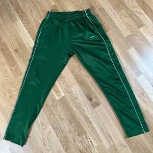 Gröna träningsbyxor Hammarby IF - Gröna träningsbyxor från Hammarby IF med vita ränder längs benen och resår i midjan. Byxorna har en sportig look och är perfekta för fotboll eller träning. Märke på ena benet. Snygga och bekväma för aktiva dagar.