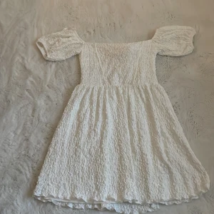 Vit klänning med puffärmar från Gina Tricot - Superfin vit klänning från Gina Tricot med puffiga kortärmar och strukturmönster i tyget. Klänningen har en söt, romantisk look med avskuren midja och lätt utställd kjol. Perfekt för sommarens tillfällen!