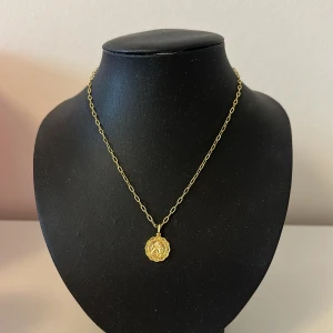 Guldigt halsband med medaljong - Snyggt halsband i guld med en rund medaljong som hänge. Kedjan är tunn och elegant, perfekt för att lyfta vilken outfit som helst. Medaljongen har ett detaljerat mönster som ger en unik touch.