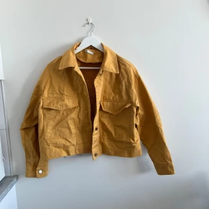 Gul croppad jacka med fickor - Säljer en gul croppad jacka med stora bröstfickor och klassisk krage. Jackan har knappar framtill och vid ärmsluten. Perfekt för dig som vill ha en färgklick i garderoben!