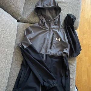 Svart och grå träningsjacka med huva från Under Armour - Säljer en snygg svart och grå träningsjacka från Under Armour med huva och dragkedja. Jackan har lång ärm, bröstficka med dragkedja och logga på bröstet. Perfekt för träning eller sportiga dagar. Väldigt eftertraktad och populär jacka!!