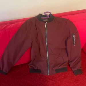 Vinröd tunn jacka  - Säljer en vinröd bomberjacka från Cometlin. Jackan har en ficka med dragkedja på ärmen och klassisk bomberkrage. Perfekt för dig som vill ha en stilren och enkel jacka.