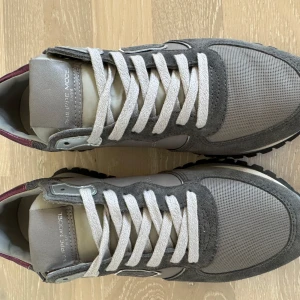 Grå Philippe Model sneakers med lila metallic-detalj - Snygga sneakers från Philippe Model i olika grå nyanser med detaljer i mocka och mesh. Skorna har en unik lila metallic-häl och vita snören. Perfekta för dig som vill ha en stilren men ändå unik look.