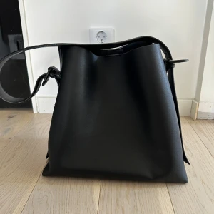 Svart axelväska i skinn - Stilren svart axelväska i slätt skinn med justerbart axelband och knytning i sidorna. Väskan har ett stort huvudfack och en innerficka med dragkedja. Perfekt för dig som vill ha en rymlig och enkel väska till vardagen.