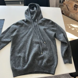 Grå hoodie i kashmir - Mysig grå hoodie i kashmir med dragsko i huvan. Tröjan har långa ärmar och ribbade muddar vid ärmslut och nederkant. Perfekt för en grishig look