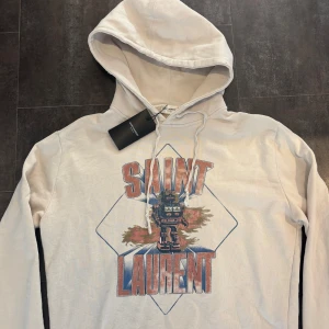 Saint Laurent Hoodie - Tja! Säljer denna riktigt feta beige/vita Saint Laurent hoodien i nyskick! Hoodien är somsagt i nyskick och har inga tecken på använding alls. Slitningarna på luvan och muddarna är design. Nypris ca 9000kr. Size S men stor i storleken så skulle säga M. Självklart äkta. Skriv vid funderingar!