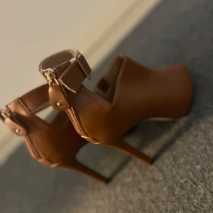Bruna klackskor med öppen tå - Snygga bruna klackskor i skinn med öppen tå och justerbart spänne runt ankeln. Perfekta för en stilren look.