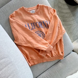 Orange sweatshirt med tryck 🧡💙 - En snygg orange sweatshirt med tryck från PULL&BEAR i storlek M men är lite liten i storleken för mig. Köpt secondhand och har använts några gånger och är lite nopprig men fortfarande i gott skick! FÖRST TILL KVARN!💙🧡(BUNDLERABATT VID KÖP AV FLER ARTIKLAR!)