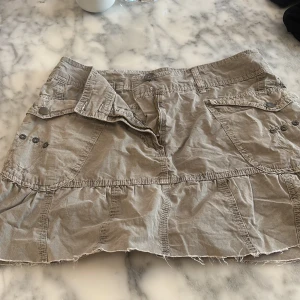 Beigegrön kortkjol med fickor - Säljer en beigegrön kortkjol med flera fickor och bälteshällor. Kjolen har en avslappnad stil med råa kanter och knappar som detaljer. Perfekt för en casual look. Saknas en knapp