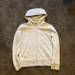Grå hoodie från GANT - Säljer en stilren grå hoodie från GANT med dragkedja och broderad logga på bröstet. Tröjan har långa ärmar och en praktisk huva. Perfekt för en avslappnad look.