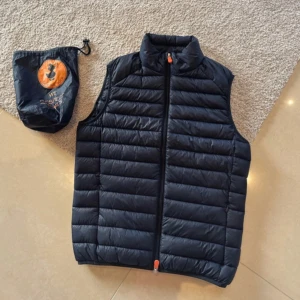 Svart dunväst från Save The Duck - Säljer en svart dunväst från Save The Duck. Västen är quiltad med dragkedja och har en praktisk förvaringspåse. Perfekt för kyliga dagar och lätt att packa med sig.