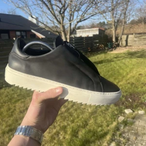 Arigato med dragkedja - Snygga svarta Arigatos i skinn med dragkedja framtill. Skorna har en vit sula med räfflad design för extra grepp. Tillverkade i Portugal, dessa sneakers kombinerar stil och funktionalitet.