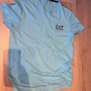 Ljusblå t-shirt från EA7 - Säljer en ljusblå t-shirt från EA7 med korta ärmar och rund hals. Perfekt för en avslappnad stil. Logotypen är tryckt på bröstet. Använd 2-3 gånger bara 