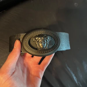 Svart Versace reversible bälte - Snyggt svart bälte med ett stort, dekorativt spänne som har ett klassiskt motiv. Perfekt för att ge en stilren touch till din outfit. Bältet är tillverkat i ett material som liknar skinn och har en elegant design.