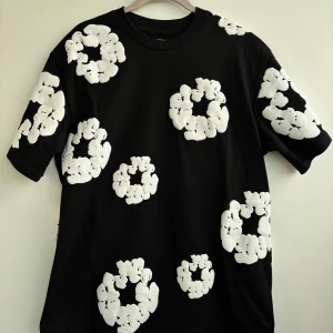 Denim Tears t-shirt  - Snygg svart t-shirt från Denim Tears med stora vita blommönster över hela plagget. Perfekt för en unik och stilren look. Kortärmad och i bekväm bomull. Condition 10/10, använt ba en gång och insåg att den är för stor för mig. Storleken är M och kan även passa någon med ett mindre L.