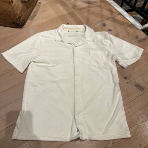 Vit kortärmad skjorta - Säljer en stilren vit kortärmad skjorta med klassisk krage och knappar framtill. Skjortan har en bröstficka och är perfekt för en avslappnad look. Materialet ser mjukt och bekvämt ut. Materialet är frotté.
