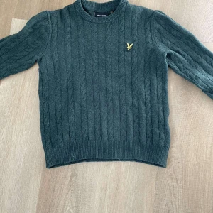Grön stickad tröja från Lyle & Scott - Säljer en snygg grön stickad tröja från Lyle & Scott med kabelstickat mönster. Tröjan har en rund halsringning och långa ärmar. Perfekt för kyligare dagar!