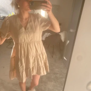 Beige kortklänning med puffärmar - Säljer en söt beige kortklänning med puffiga kortärmar och v-ringning. Klänningen har en luftig passform och volangdetaljer som ger en romantisk känsla. Perfekt för varma sommardagar!