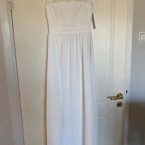 Vit långklänning med spetsdetaljer - Elegant vit långklänning med vackra spetsdetaljer och blommönster. Klänningen har en romantisk känsla med sin ärmlösa design och långa fall. Perfekt för speciella tillfällen.