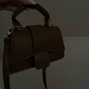 Brun handväska med axelrem - Snygg brun handväska i skinn med en praktisk axelrem och ett elegant handtag. Väskan har en stilren design med ett lock och spänne framtill. Aldrig använd 