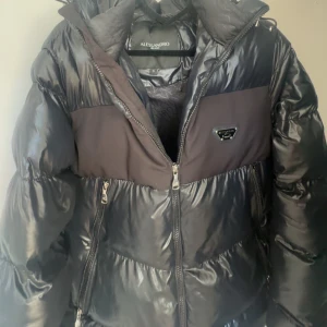 Svart pufferjacka från Alessandro - Snygg svart pufferjacka från Alessandro med glansig finish och dragkedja framtill. Jackan har en hög krage och huva för extra värme. Perfekt för kyliga dagar.