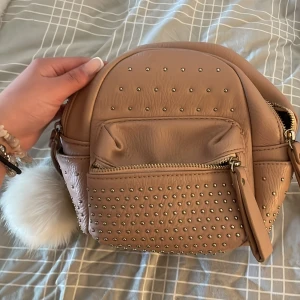 Beige axelväska med nitar - Säljer en snygg beige axelväska med nitar och dragkedja. Väskan har en fluffig vit boll som dekoration och praktiska handtag. Perfekt för att ge din outfit en cool touch!