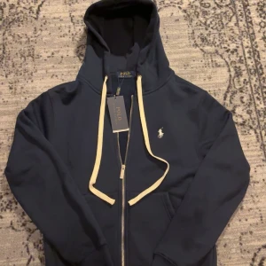 Ralph Lauren Zip Hoodie - Säljer en snygg mörkblå hoodie från Ralph Lauren med dragkedja och vit logga på bröstet. Den har en justerbar huva med vita snören och ribbade muddar vid ärmslut och nederkant. Perfekt för en stilren och bekväm look. Helt ny med prislapp kvar, skick 10/10, pris kan diskuteras vid snabbaffär! Frågor? DM!