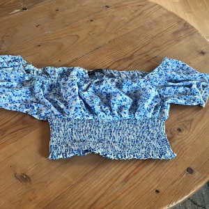 Blå blommig offshoulder topp - Säljer en söt blå offshoulder topp med blommigt mönster. Toppen har en elastisk smockdetalj vid midjan och korta ärmar. 