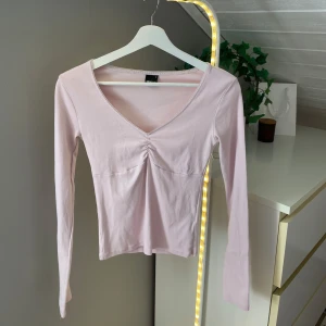 Ljusrosa långärmad topp Gina Tricot - Supersöt ljusrosa långärmad topp från Gina Tricot med v-ringning och rynkdetalj framtill. Toppen är figurnära och i mjuk bomull. Tröjan är använd max 5 gånger men har tyvärr lossat en liten tråd i armhålan!