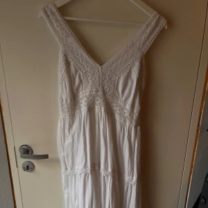 Vit långklänning från Corte Hindu -  vit långklänning från Corte Hindu med v-ringning, breda axelband och vackra spetsdetaljer. Klänningen har omlottkänsla upptill, smock i ryggen och volanger längs kjolen. Perfekt för sommarens alla tillfällen. Ungefär storlek M