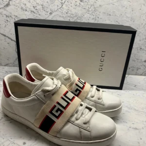 Gucci Stripe Sneakers  - Snygga vita sneakers från Gucci med röd och svart logotyprem över snörningen. Skorna har en röd detalj på hälen och är tillverkade i skinn. De är i storlek 45 men sitter som 44. Perfekta för alla säsonger. Du får med boxen, extra snören och authority card när du köper skorna. 