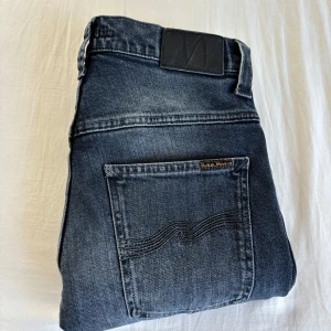 Jeans från Nudie Jeans Co - Snygga blå jeans från Nudie Jeans Co med klassisk femficksdesign och diskret logga på bakfickan. Jeansen har raka ben och normal passform, tillverkade i slitstarkt bomullsjeans. Perfekta för dig som gillar stilrena och tidlösa jeans.