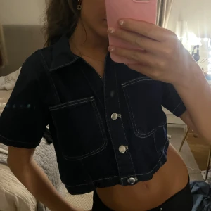 Mörkblå croppad jeansskjorta XS - Snygg croppad skjorta i mörkblå denim med korta ärmar och stora bröstfickor. Skjortan har silvriga knappar framtill och markerade sömmar som ger en cool look. Perfekt för dig som gillar streetstyle och vill sticka ut lite extra.