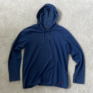 Marinblå hoodie från Polo Ralph Lauren - Snygg marinblå hoodie från Polo Ralph Lauren med klassisk huva och dragsnören. Tröjan har lång ärm och är tillverkad i mjuk bomull. Liten röd broderad logga nertill på framsidan ger en stilren touch. Perfekt för en chill och avslappnad look.