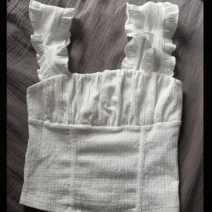 Vit volangtopp med breda axelband - Supersöt vit topp med breda volangprydda axelband och smockad struktur. Toppen är croppad och har en lätt figurnära passform. Perfekt för varma dagar och ger en romantisk vibe till din outfit.