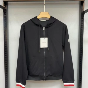 Moncler zip-up hoodie - Svart vindjacka från Moncler med huva, dragkedja framtill och fickor. Ärmsluten har vita och röda detaljer för en sportig look. Klassisk Moncler-logga på ärmen. Perfekt för dig som gillar stilren design och streetwear-vibe.