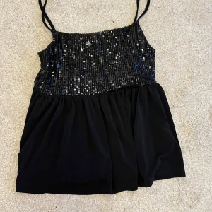 Svart paljettlinne med tunna band - Supersnygg svart linne-topp med tunna axelband och glittrig paljettöverdel. Toppen har en flowy, luftig nederdel och passar perfekt när du vill sticka ut. Paljetterna är i svart och silver med inslag av blått för extra effekt.