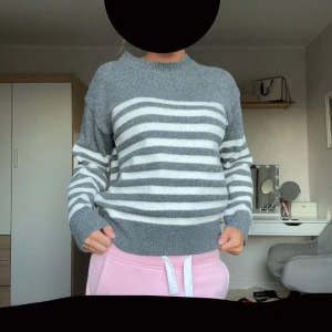 Randig grå och vit stickad tröja - Supermysig stickad tröja i grått och vitt med horisontella ränder över bröstet och ärmarna. Tröjan har en rund halsringning och långa ärmar med breda muddar. Perfekt för kyliga dagar och enkel att matcha med jeans eller mjukisbyxor.