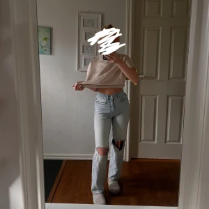 Ljusblå ripped jeans med raka ben - Säljer ett par ljusblå jeans med raka ben och stora slitningar vid knäna. Byxorna har hög midja och klassisk femficksdesign. Perfekta för en avslappnad och trendig look.
