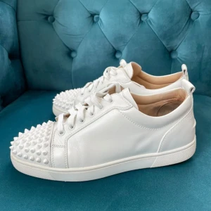 Vita Louboutin sneakers med nitar - Snygga vita sneakers från Christian Louboutin i skinn med ikoniska röda sulor och coola nitar på tån. Skorna har snörning och en klassisk låg siluett som sticker ut med sina detaljer. Perfekta för dig som vill ha något extra till din outfit.