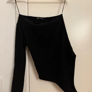 Svart one shoulder body från Zara - Säljer en svart one shoulder body från Zara med lång ärm på ena sidan och smal axelrem på andra. Snygg asymmetrisk design som ger en cool och trendig look. Tunn och mjuk i materialet, perfekt att styla med jeans eller kjol.