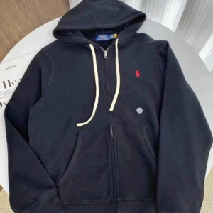 Svart hoodie från Polo Ralph Lauren - Svart hoodie från Polo Ralph Lauren med vit dragsko och dragkedja framtill. Klassisk röd logga broderad på bröstet och två stora fickor. Perfekt för en chill och stilren look.