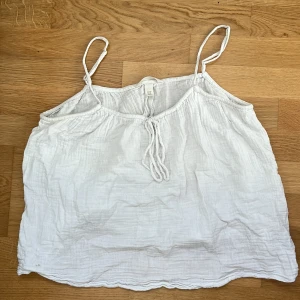 Vit linnetopp med knyt från H&M - Superfin vit linnetopp från H&M med tunna axelband och knyt framtill. Toppen är gjord i ett luftigt bomullstyg med lätt skrynklig struktur, perfekt för varma dagar. Enkel och stilren design som är lätt att matcha. 