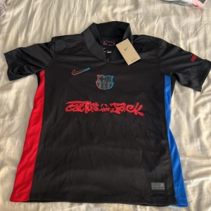 FC Barcelona x Nike Lamine Yamal tröja - Snygg svart FC Barcelona fotbollströja från Nike med Lamine Yamal och nummer 19 på ryggen. Tröjan har röda och blå detaljer på sidorna, klubbmärke på bröstet och coolt Cactus Jack-tryck framtill. Tillverkad i Dri-FIT-material som andas, perfekt för match eller träning.