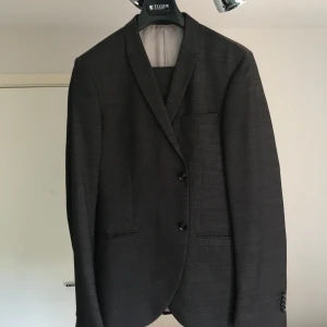 Mörkgrå kostym set från Tiger of Sweden - Snygg mörkgrå kostym set från Tiger of Sweden med klassisk kavaj och matchande byxor. Kavajen har två knappar, smala slag och innerfoder i ljusgrått. Perfekt för dig som gillar stilren och tidlös design.