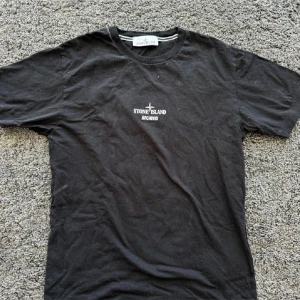 Svart Stone Island Archivio t-shirt S - Svart t-shirt från Stone Island Archivio med vit logga tryckt på bröstet och en stor röd grafik på ryggen. Klassisk rund hals och korta ärmar. Tillverkad i mjuk bomull, perfekt för dig som gillar streetwear med en unik touch. Aldrig använd, köpt på NK i Stockholm. Köpte för 2200kr. Äkta✅
