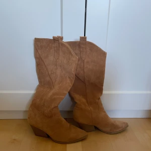 Beiga mocka cowboy boots med klack - Snygga cowboy boots i beige mocka med spetsig tå och markerad klack. Klassisk westernstil med högt skaft och dragflikar upptill. Perfekta för dig som vill sticka ut med en trendig och tidlös look.