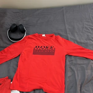 Röd långärmad tröja från Hugo Boss - Snygg röd långärmad tröja från Hugo Boss med svart logga på bröstet. Tröjan har en klassisk passform och är gjord i mjuk bomull, perfekt för dig som gillar stilrena och bekväma plagg. Passar till jeans eller joggers för en chill look.