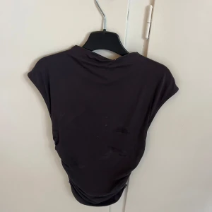 Svart croppad topp med rynkade sidor - Säljer en svart croppad topp med ärmlös design och snygga rynkade detaljer längs sidorna. Toppens material är mjukt och stretchigt, perfekt för en trendig och avslappnad look. Stl xxs men är väldigt stretchig så passar nog xs